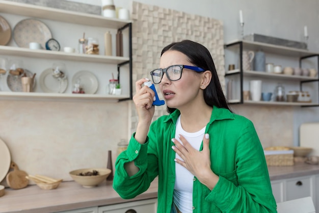 Eine junge Frau, die an Asthmaanfällen leidet, benutzt zu Hause in der Küche einen Inhalator und hält sich dabei die Brust