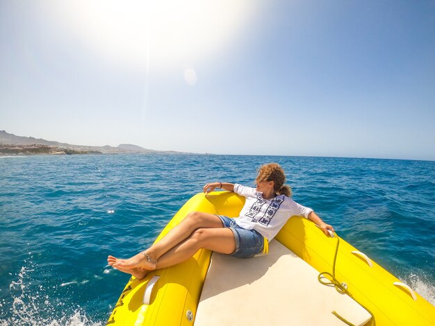 Eine junge Frau, die auf einem Boot liegt und das Meer entdeckt und einen sonnigen Tag genießt und sich entspannt - mit Blick auf die Küste vom Beiboot aus