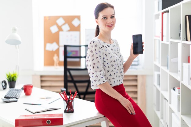 Eine junge Frau, die im Büro steht und ein Telefon hält.