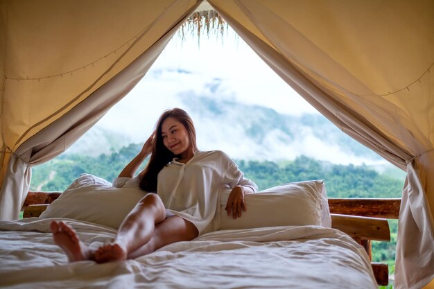 Eine junge Frau, die sich morgens auf einem weißen Bett mit einem wunderschönen Blick auf die Natur außerhalb des Zeltes hinlegt