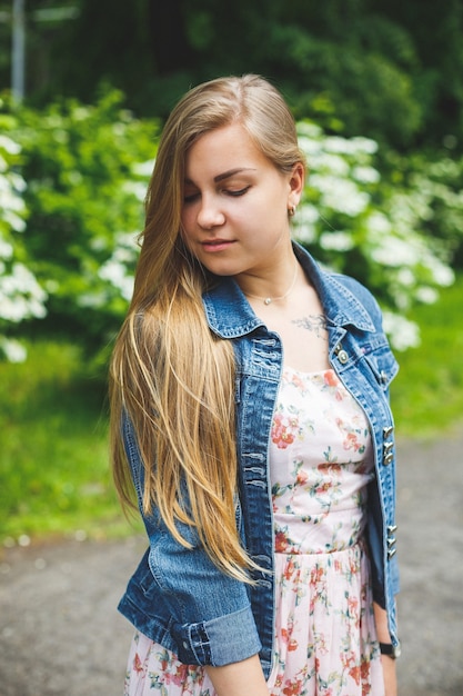 Eine junge Frau europäischen Aussehens mit langen blonden Haaren, gekleidet in ein kurzes Kleid, steht vor dem Hintergrund weiß blühender Büsche. Sonniger Frühlingstag. Natürliche weibliche Schönheit