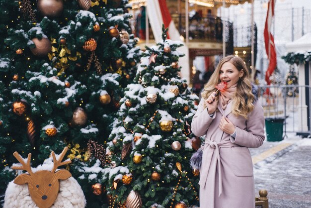 Eine junge Frau geht zu Weihnachten auf dem Platz in der Nähe der geschmückten Weihnachtsbäume. Candy ist ein Lutscher in Form eines Herzens.