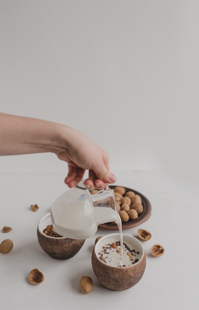 Eine junge Frau gießt Milch in eine Schüssel mit Müsli. Eine Hand hält einen Milchkännchen, einen Krug Milch. Ein gesundes Frühstück aus Müsli, Haferflocken, Müsli, Nüssen.