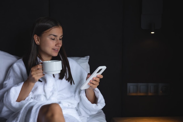 Eine junge Frau im Bademantel mit einer Tasse Kaffee und einem Smartphone im Bett