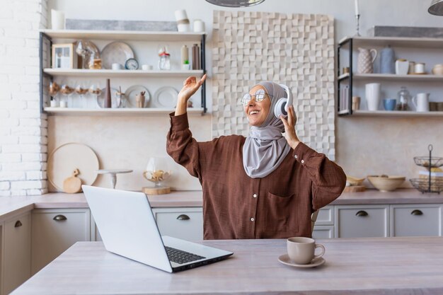 Eine junge Frau im Hijab orientalischer Herkunft sitzt zu Hause in der Küche und bedient einen Laptop