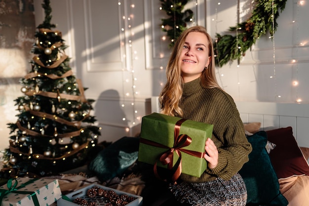 Eine junge Frau im Pullover öffnet Weihnachtsgeschenke in einem weihnachtlich dekorierten Schlafzimmer vor der Kulisse eines Weihnachtsbaumes.