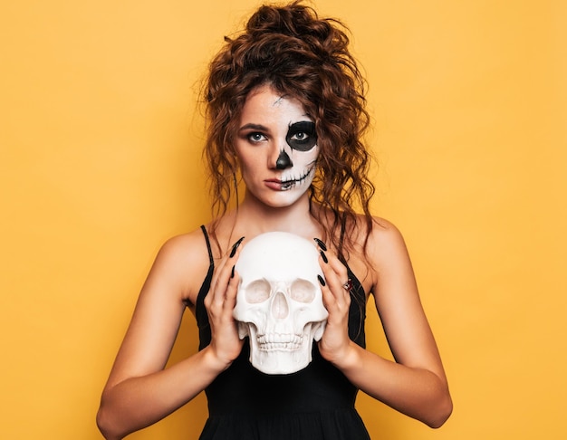 Eine junge Frau in einem Halloween-Kostüm mit Make-up für den Urlaub auf einem gelb isolierten Hintergrund mit einem Totenkopf in den Händen Die Emotion im Gesicht ist beängstigend