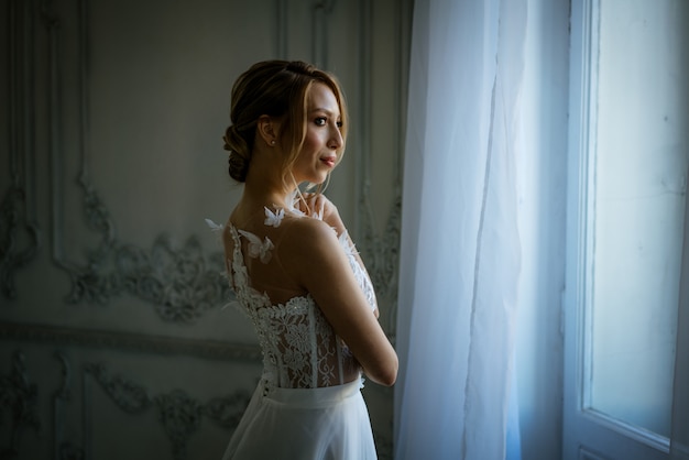 Eine junge Frau in einem Hochzeitskleid und einer Frisur steht am Fenster, eine schöne Silhouette