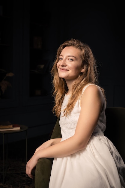 Eine junge Frau in einem Kleid mit langen Haaren lächelt und schaut in einem dunklen Raum in die Kamera