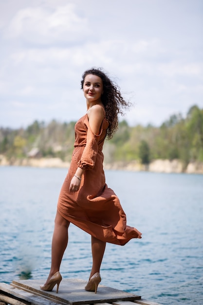 Eine junge Frau in einem Kleid steht auf einem Holzmauerwerk inmitten eines blauen Sees. Mädchen lächelt glücklich und die Sonne scheint, Sommertag. Sie hat lockiges langes Haar und ein europäisches Aussehen
