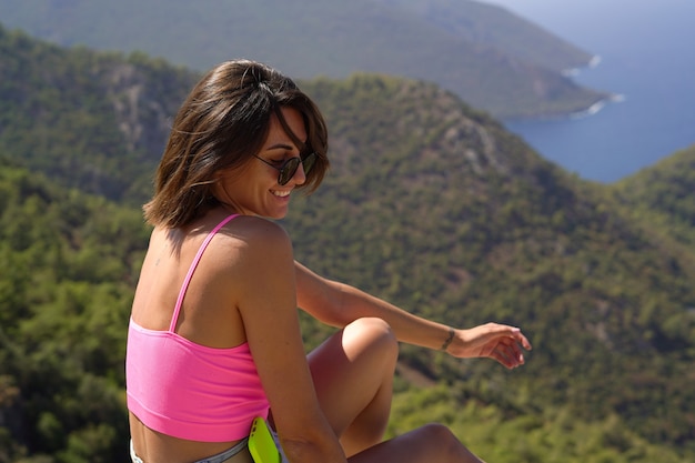 Eine junge Frau in einem leuchtend rosa Top sitzt auf einem Berggipfel mit herrlichem Blick auf die Berge und das Mittelmeer im Süden der Türkei