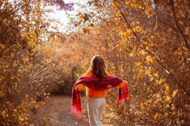 Eine junge Frau in einem orangefarbenen Pullover hüllt sich in einem Herbstpark in einen Schal