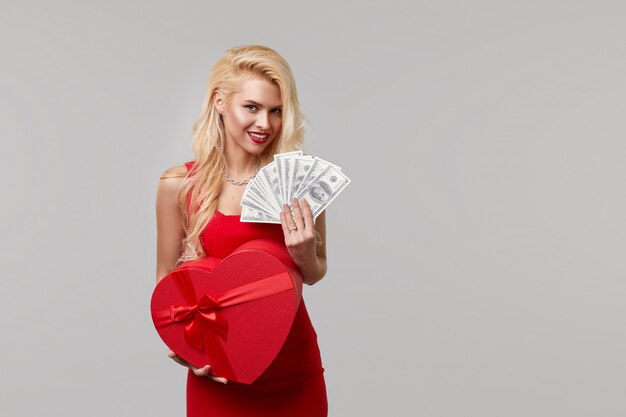 Eine junge Frau in einem roten Kleid hält in ihren Händen Bargeld, Geld, Dollar und eine herzförmige Geschenkbox. Valentinstag und 8. März
