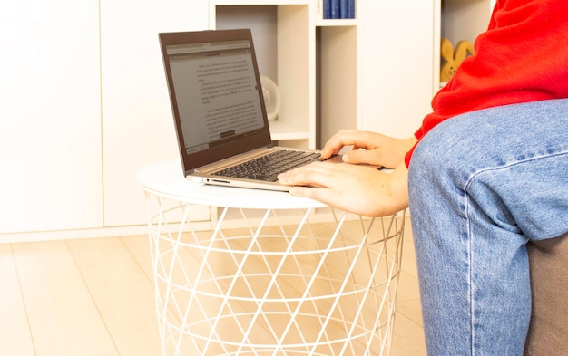 Eine junge Frau in einem roten Pullover sitzt im Zimmer und arbeitet zu Hause mit einem Laptop