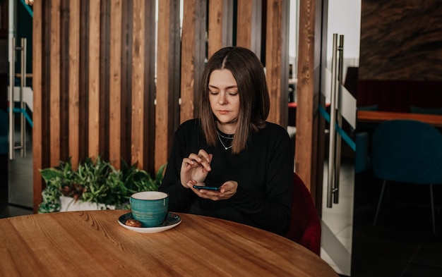 Eine junge Frau in einem schwarzen Kleid sitzt mit einer Tasse Kaffee und einem Telefon an einem Tisch in einem Café