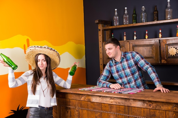 Eine junge Frau in einem Sombrero, die zwei Flaschen Bier hält und an der Bar neben dem Barkeeper in einem karierten Hemd in einer mexikanischen Bar steht