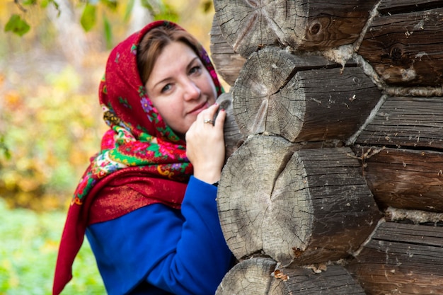 Eine junge Frau in einem traditionellen russischen Kopftuch schaut um die Ecke eines Holzhauses. Rotes Taschentuch von Pawlowsk