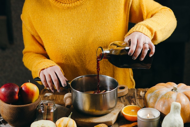 Eine junge Frau in einem warmen, gestrickten, gelben Pullover gießt Wein aus einer Glasflasche in eine Pfanne, um heißen Glühwein herzustellen