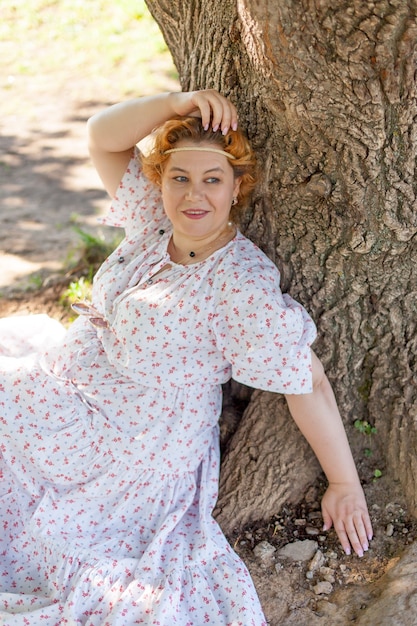 Eine junge Frau in einem weißen Kleid, sitzend auf dem Gras neben einem Baum im Freien, romantisches Sommerfoto.