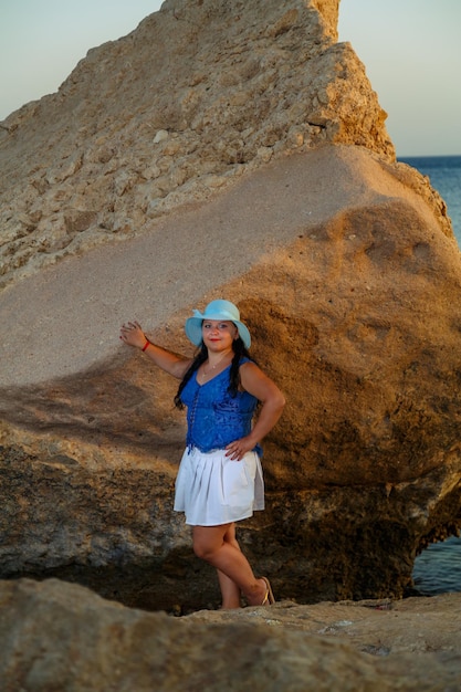 Eine junge Frau in einem weißen Rock und einem Sonnenhut steht am Ufer zwischen den Felsen vor dem Hintergrund des Meeres bei Sonnenuntergang.