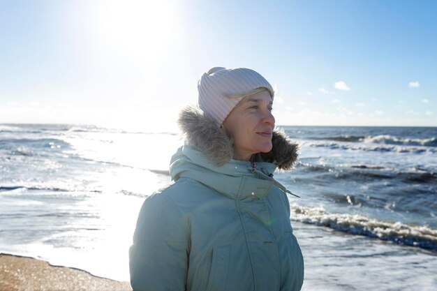 Eine junge Frau in einer blauen Jacke geht an einem sonnigen Tag in der Wintersaison entlang des Meeres. Aktiver Lebensstil und Spaziergänge in der Natur. Nahaufnahme