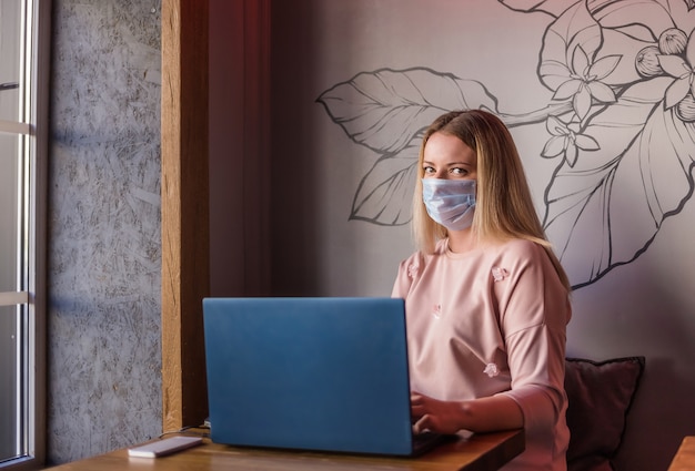 Eine junge Frau in einer Maske sitzt mit einem Laptop und einem Telefon in einem Café