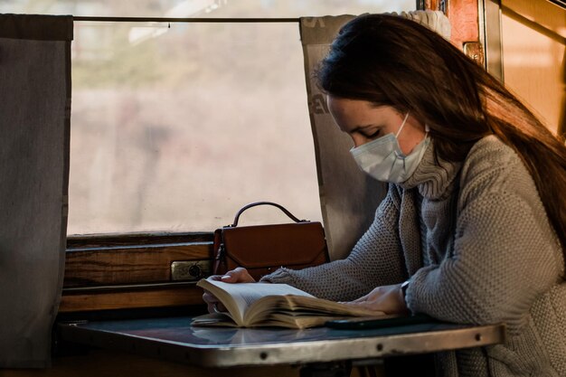 Eine junge Frau in einer medizinischen Maske fährt während einer Coronavirus-Pandemie mit dem Zug Das Mädchen liest ein Buch und hört Musik im Zug Vorsichtsmaßnahmen im öffentlichen Verkehr treffen