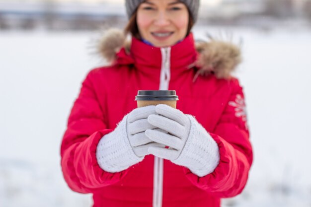 Eine junge Frau in einer roten Jacke im Winter hält ein Glas heißen Kaffee oder Tee