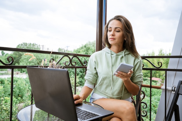 Eine junge Frau in Hauskleidung sitzt auf dem Balkon, arbeitet an einem Tisch und arbeitet am Sommernachmittag mit einem Telefon an einem Laptop