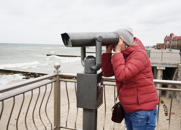 Eine junge frau in jacke und jeans schaut durch ein öffentliches teleskop am wasser und beobachtet ein wunderschönes panorama der ostsee