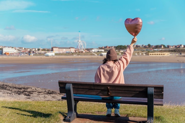 Eine junge Frau in Jeans und einer rosa Jacke, die auf einer Bank sitzt und einen Ballon an der Küste hält