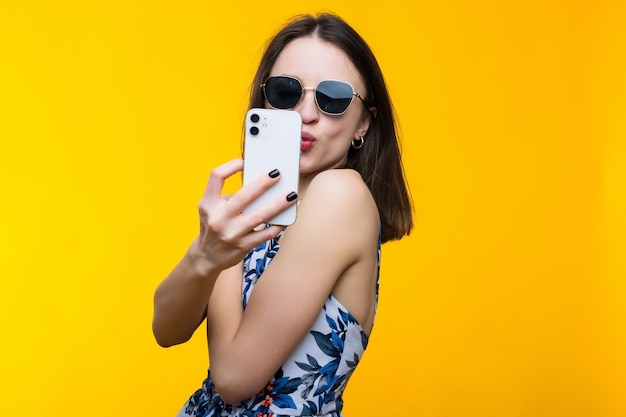 Eine junge Frau in Kleid und Sonnenbrille macht auf gelbem Hintergrund ein Selfie mit ihrem Handy