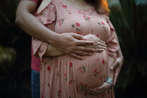 Eine junge Frau in rosa bedrucktem, eng anliegendem Kleid umarmt ihren Bauch mit einem zukünftigen Baby.