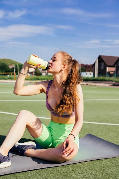Eine junge Frau in Sportklamotten sitzt auf einer Matte und trinkt Wasser