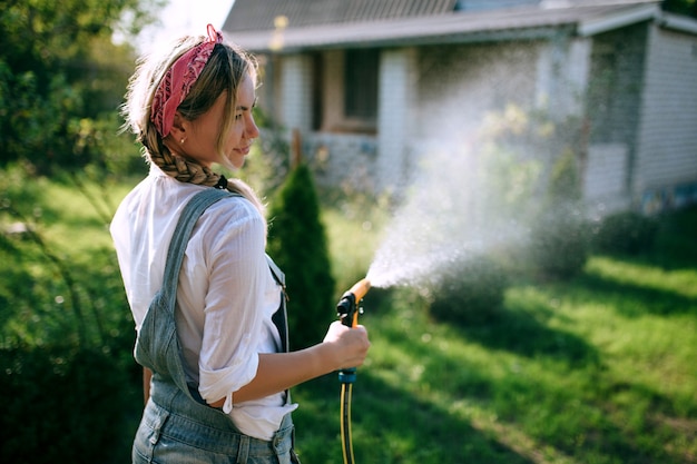 eine junge Frau in weißem Hemd und Jeans-Overall, die den Rasen bewässert