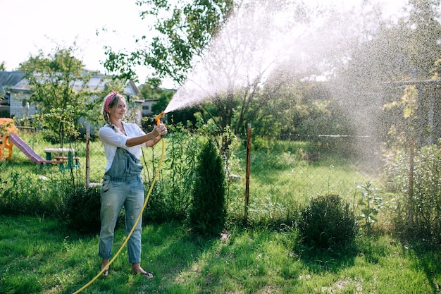 eine junge Frau in weißem Hemd und Jeans-Overall, die den Rasen bewässert