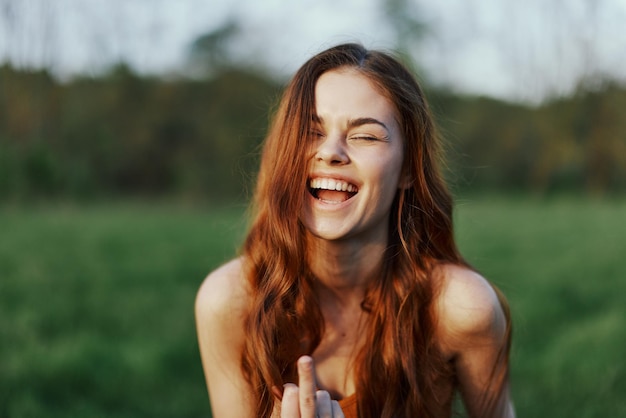 Eine junge Frau lächelt und schaut mit ihrem langen, roten, welligen, glänzenden Haar in einem Park mit grünem Gras im Sommersonnenuntergang in die Kamera Das Konzept der gesunden Lebensschönheit und Haarpflege