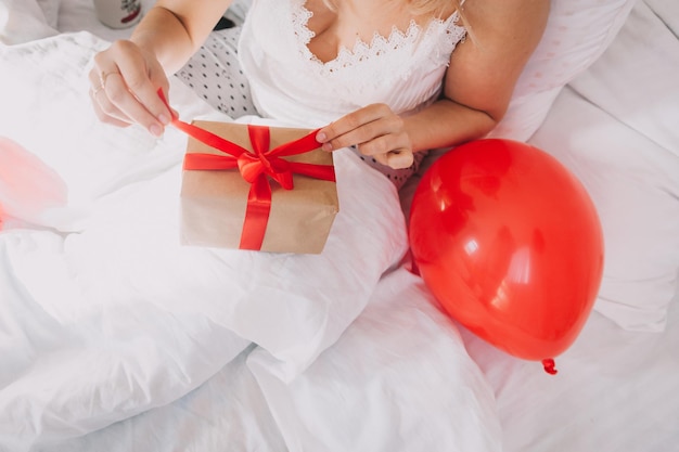 Eine junge Frau liegt auf dem Bett und öffnet eine Schachtel mit einem Geschenk. Feier des Valentinstags.