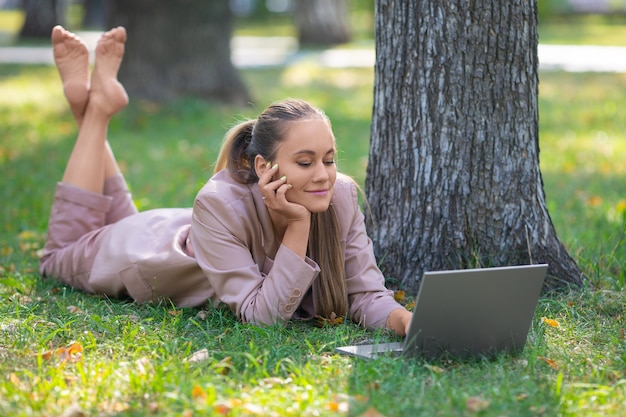 Eine junge Frau liegt im Park auf der Wiese vor einem Laptop-Monitor, den sie während einer Online-Besprechung lächelt.