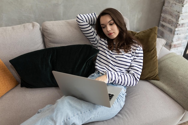Eine junge Frau liegt mit einem Laptop auf dem Sofa und schaut sich etwas im Internet an