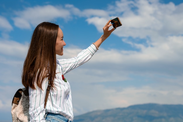 Eine junge Frau macht ein Selfie auf einem Smartphone gegen einen blauen Himmel mit Wolken