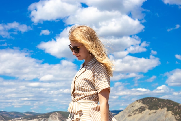 Eine junge Frau mit blonden Haaren und Sonnenbrillen neigte ihren Kopf gegen einen blauen Himmel mit Wolken in einer bergigen Gegend