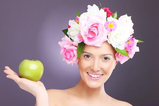 eine junge Frau mit Blumen im Haar posiert mit einem Apfel auf violettem Hintergrund