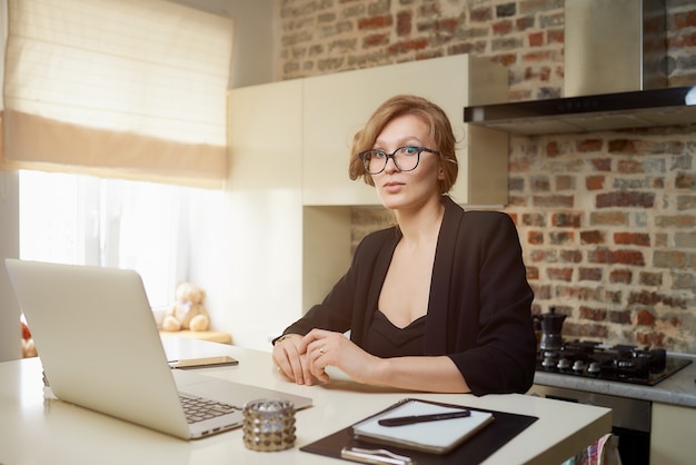 Eine junge Frau mit Brille arbeitet in ihrer Küche an einem Laptop. Ein blondes Mädchen hört ihren Kollegen auf einer Videokonferenz zu Hause zu. Eine Dame, die eine Lektion online in einem Webinar lernt.