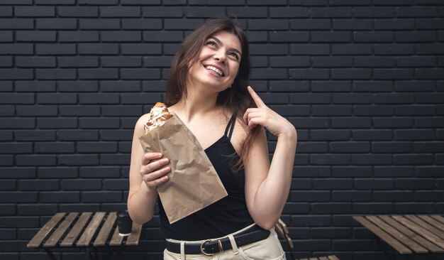 Eine junge Frau mit einem Croissant und einer Tasse Kaffee vor einer schwarzen Backsteinmauer