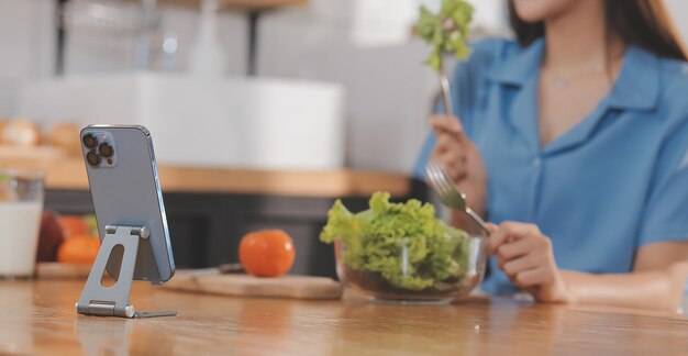 Eine junge Frau mit einem schönen Gesicht in einem blauen Hemd mit langen Haaren, die Obst isst, sitzt zu Hause in der Küche mit einem Laptop und einem Notebook zum Entspannen Konzepturlaub