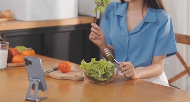 Eine junge Frau mit einem schönen Gesicht in einem blauen Hemd mit langen Haaren, die Obst isst, sitzt zu Hause in der Küche mit einem Laptop und einem Notebook zum Entspannen Konzepturlaub