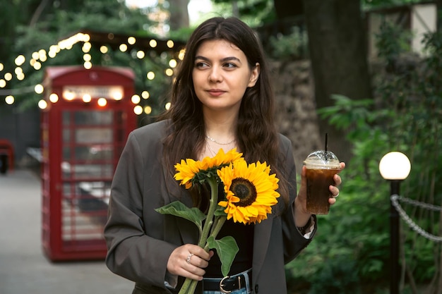 Eine junge Frau mit einem Strauß Sonnenblumen und einem kalten Kaffeegetränk in der Stadt