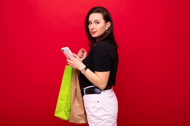 Eine junge Frau mit einer Einkaufstasche und einem Telefon in der Hand auf rotem Hintergrund kauft im Einkaufszentrum Geld ein