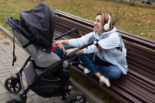 Eine junge Frau mit Kinderwagen im Park hört auf einer Bank sitzend Musik über Kopfhörer